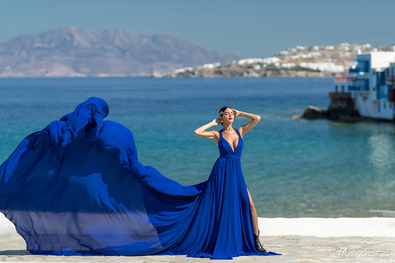 Woman in a royal blue flying dress with a deep v-neckline posing in Little Venice, Mykonos, with the dress billowing dramatically against the turquoise sea and island backdrop.